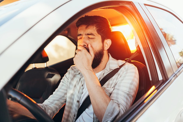 Man driving a car at sunset while yawning and covering his mouth with his hand.