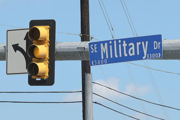 Traffic light at SE Military Drive intersection with the yellow light illuminated in the center lens, next to a left-turn sign and street sign, set against a clear blue sky with light clouds and overhead utility wires.