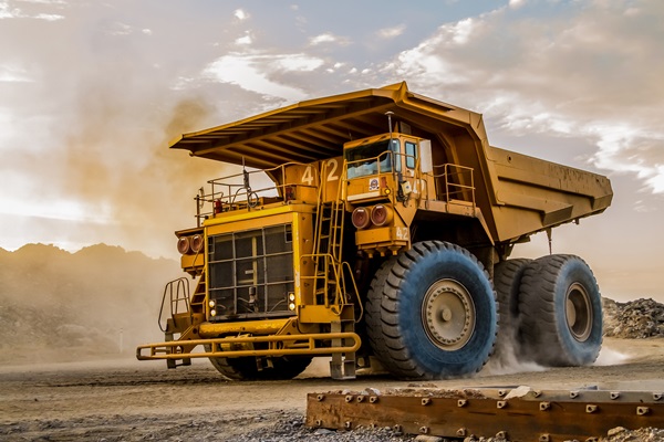 A massive, yellow dump truck with oversized blue-rimmed tires driving on a dirt road near a construction site, kicking up dust against a cloudy sunset sky.