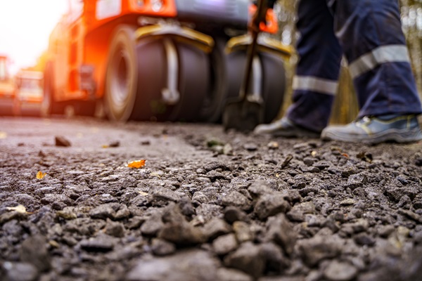 Freshly laid asphalt and loose gravel in the foreground, with the lower legs of a construction worker and the bright orange steamroller used for paving visible behind in the soft afternoon light.