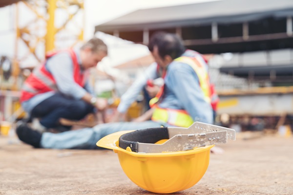 A yellow hard hat rests in the foreground of a Texas construction site while blurred figures of workers assist an injured colleague in the background.