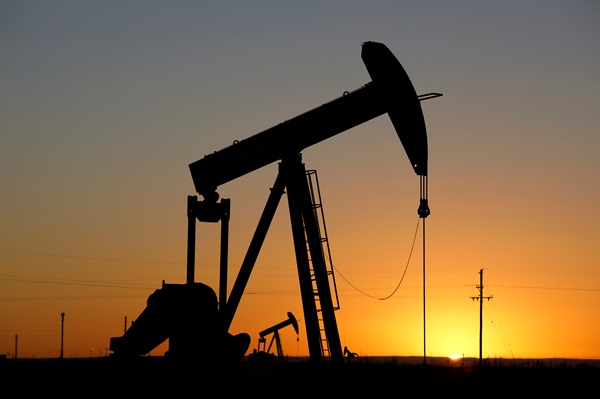A silhouette of a large oil pump jack against a vibrant orange and yellow sunset. Several smaller oil rigs are visible in the distance across a flat horizon under a clear sky in San Antonio, TX.