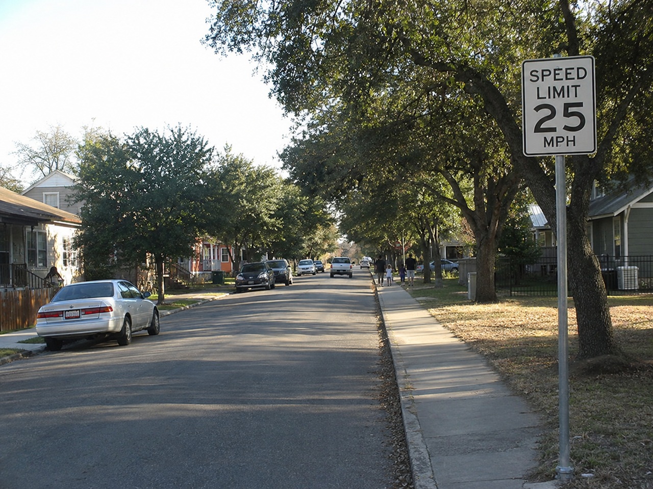 A quiet, tree-lined residential street features a prominent "SPEED LIMIT 25 MPH" road sign in the foreground, with a silver sedan parked on the left and pedestrians walking on the sidewalk in the distance, representing neighborhood traffic safety, municipal speed regulations, and car accident personal injury claims in San Antonio, Texas.