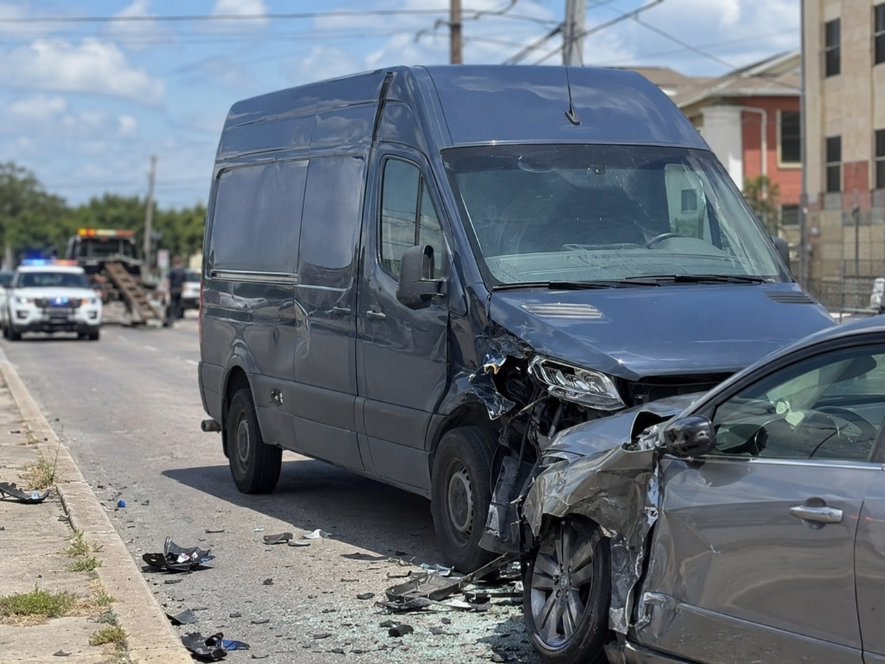 A gray Amazon-style delivery sprinter van sits nose-to-nose with a heavily damaged sedan on a Texas street, with crushed front ends and debris scattered across the pavement and a police vehicle and tow truck visible in the background, depicting a collision involving a last-mile delivery van that may give rise to a lawsuit against Amazon, its delivery service partner, or both under Texas law.