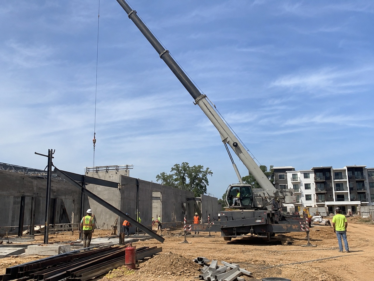 A large mobile crane extends its boom over an active Texas construction site where workers in high-visibility vests and hard hats direct a lift near partially erected concrete tilt-wall panels and steel beams, with a residential apartment complex visible in the background, depicting the heavy equipment operations and multi-contractor environment where crane accidents and serious worker injuries can give rise to liability claims against crane operators, contractors, and site owners under Texas law.
